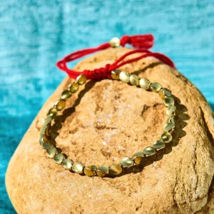 Handcrafted copper bead bracelet with red tassel closure displayed on stone background, matte gold finish for Buddhist protection and prosperity