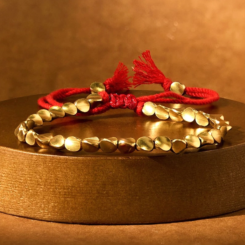Two stacked matching copper bead bracelets with red tassel closures on wooden pedestal, shiny metallic finish for spiritual abundance