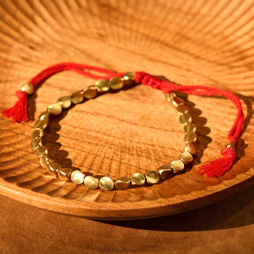 Flat copper bead bracelet with dual red tassels on carved wooden plate, traditional Buddhist design with woven central knot detail
