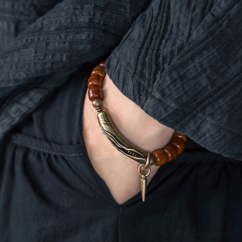 Buddhist protection bracelet styled under dark linen sleeve, oblique wrist view displaying antique bronze metal clasp, reddish barrel beads, and spike charm tucked beneath fabric