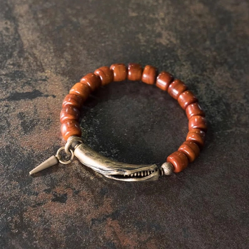 Buddhist strength bracelet with reddish barrel beads and bronze clasp featuring spike pendant, angled top-down view on textured dark background