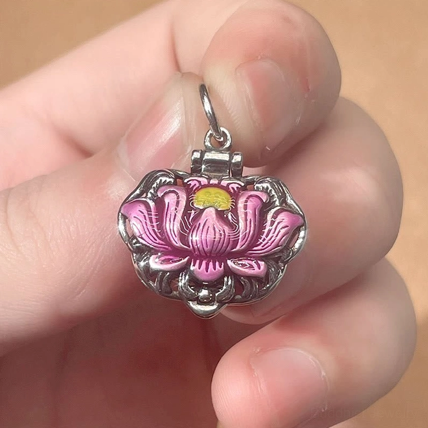 Close-up detail of copper lotus pendant held in fingertips, showing hinged bail, pink enamel lotus with yellow center and fine craftsmanship