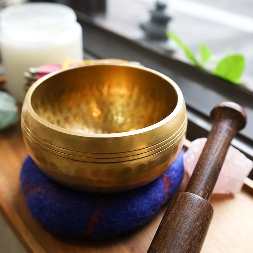 Hand-hammered copper singing bowl on felt cushion with wooden striker on meditation windowsill, close-up view of crafted texture