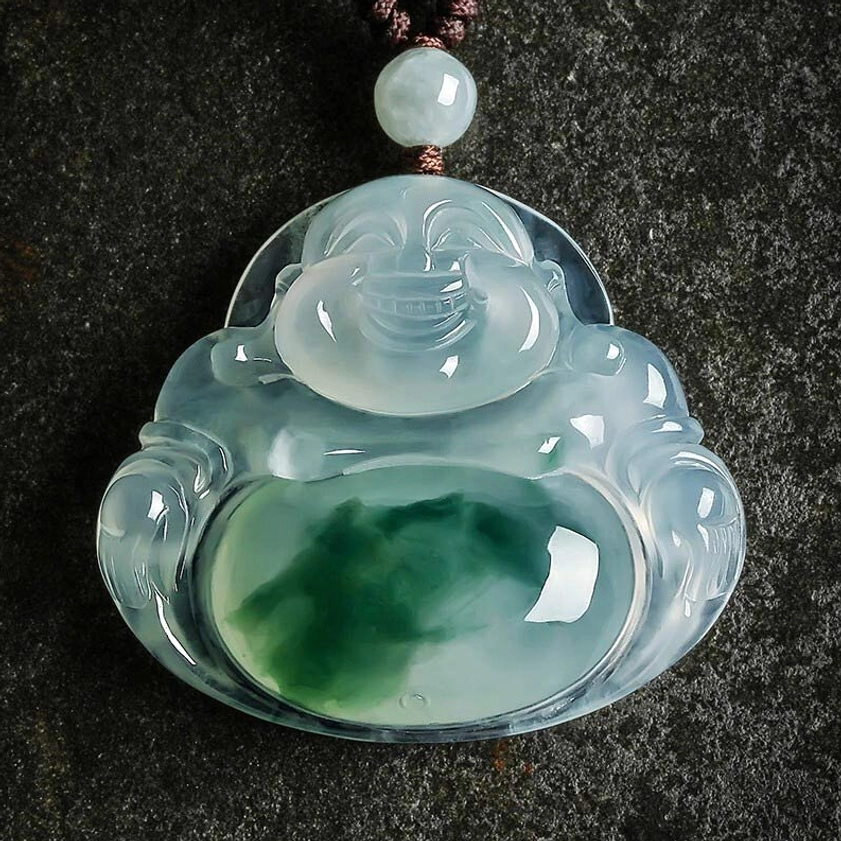 Handcrafted cyan jade laughing Buddha pendant with carved smiling Buddha face and dark green jade veining, displayed on dark stone background with brown waxed cord