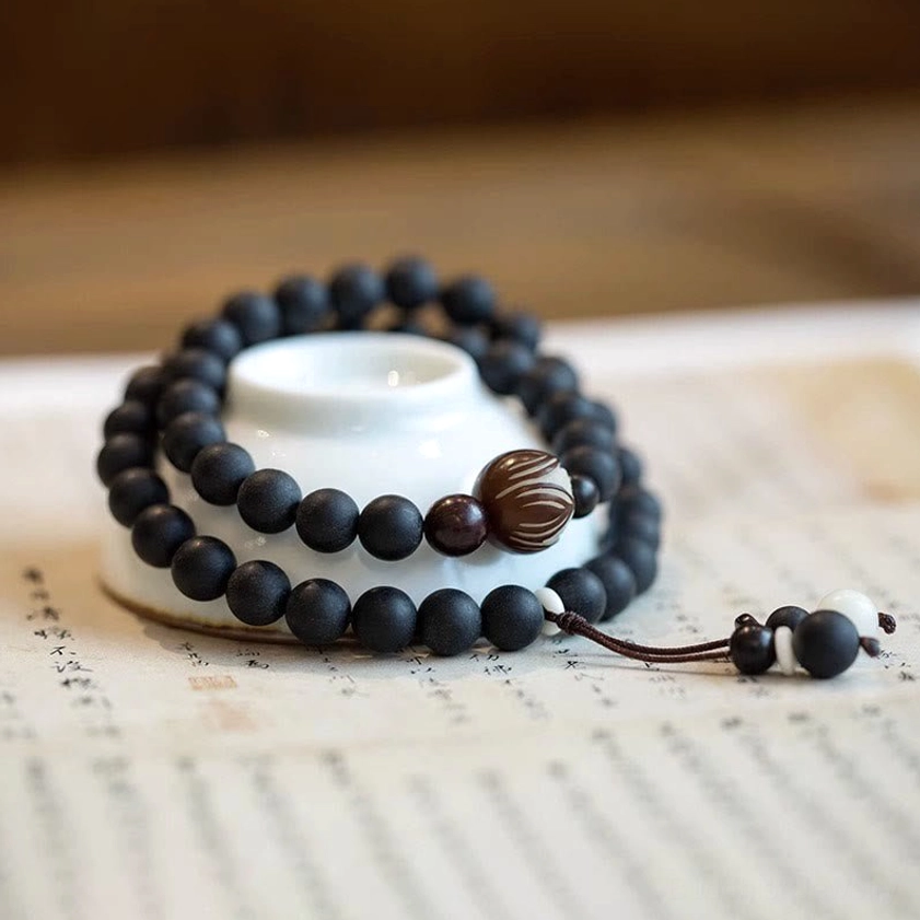 Cypress wood prayer bracelet with carved lotus bodhi bead and black tassel displayed on vintage paper with porcelain tea cup