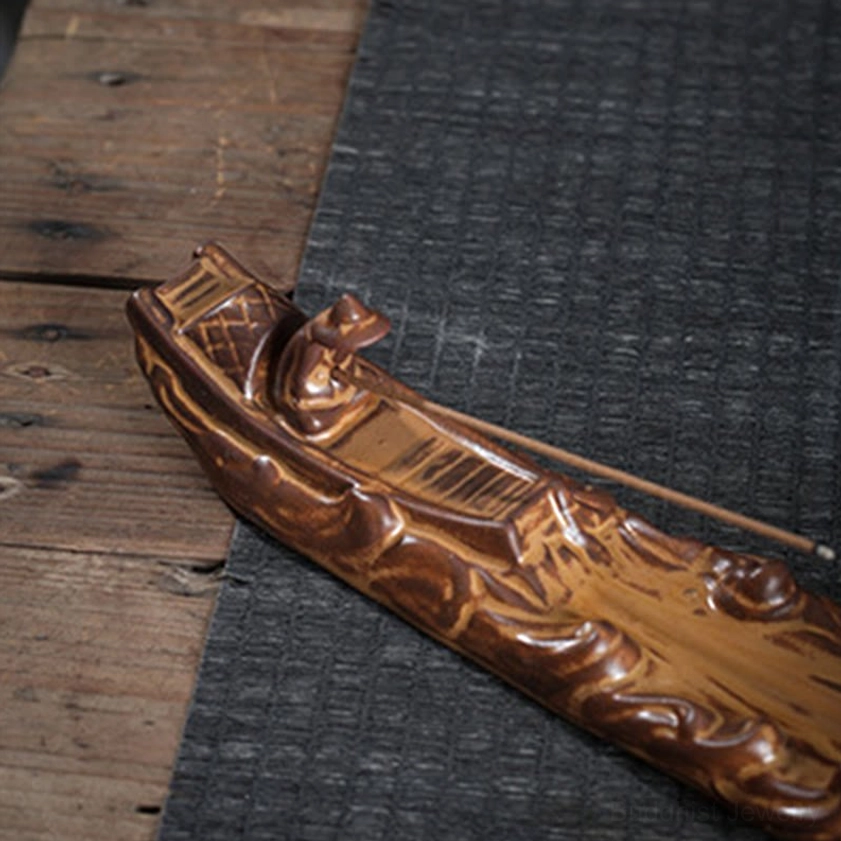 Boat-shaped ceramic incense burner with detailed deck carving and wavy hull texture on wooden table with woven mat