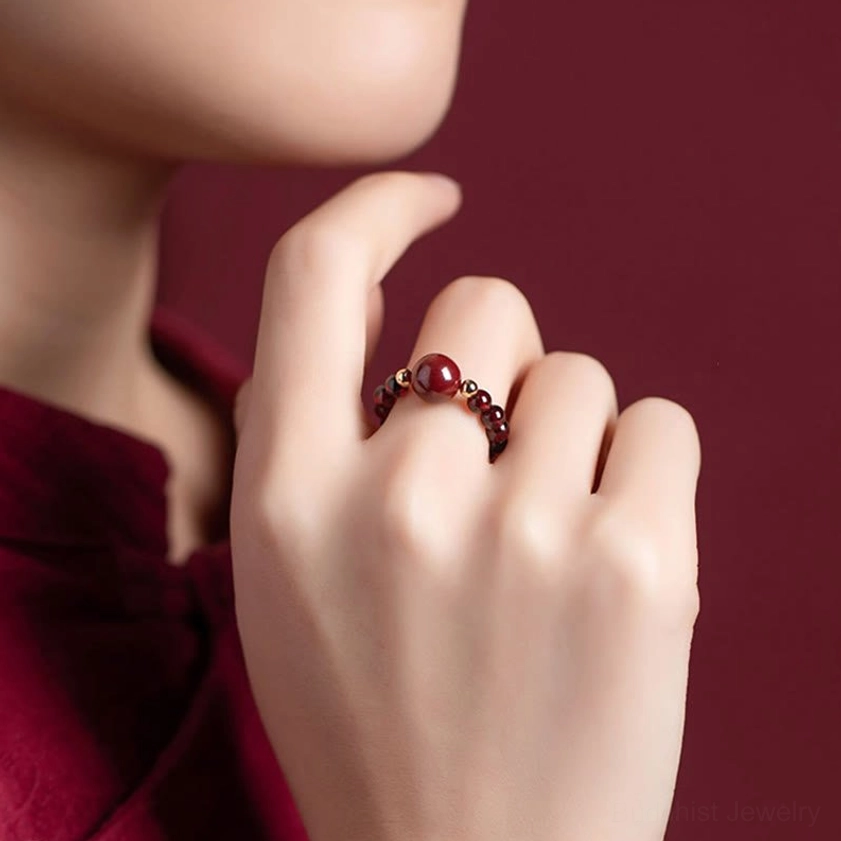 Close-up of delicate cinnabar beaded Buddhist ring with small red cinnabar accent detail worn on middle finger against wine-red clothing, showing fine craftsmanship