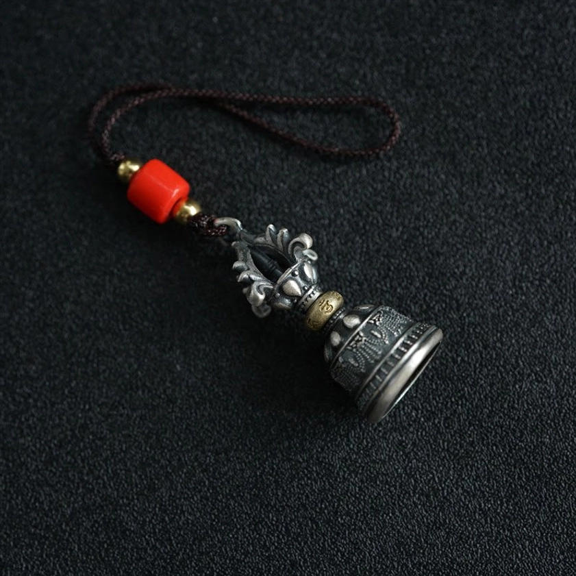 Tibetan dorje bell hanging decoration side view with red bead accent and brown decorative cord on fabric background