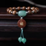 Abelia Biflora dragon wood Buddhist bracelet with 15 beads, green jade, Fu charm, and decorative tassel on dark wooden surface - top-down flat lay view