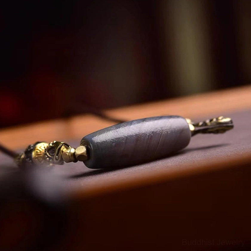 Polished grey banded agate cylinder Dzi bead with metal end fittings and woven cord detail on wooden surface