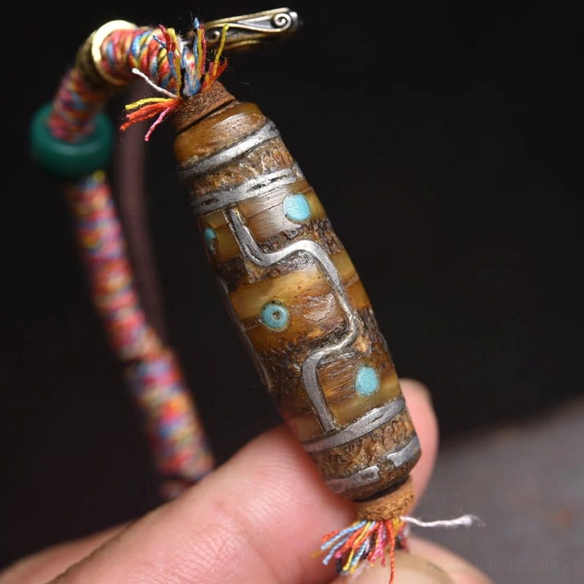 Close-up handheld view of nine-eye dzi bead with brown stone base, silver patterns and turquoise inlays paired with colorful braided rope
