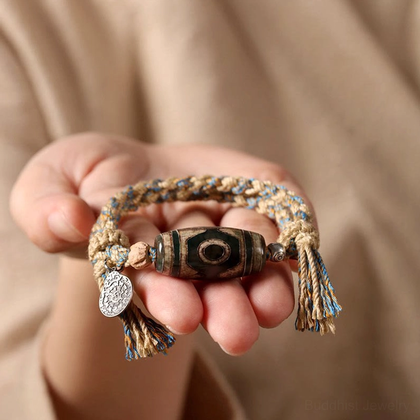 Close-up of dzi bead protection bracelet held in palm showing beige-blue woven detail and silver charm