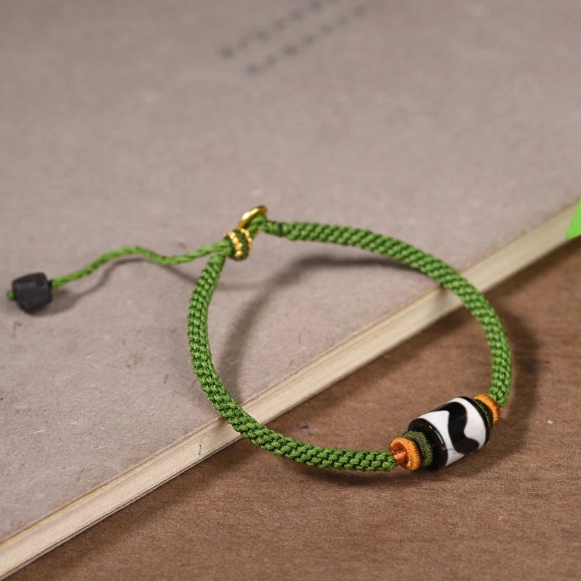 Side flat lay of handmade dzi bead bracelet with green braided cord and orange accent knots on wooden surface