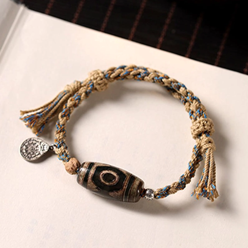 Handmade Buddhist Dzi bead string bracelet with engraved copper alloy tag detail on vintage rope, flat lay product shot