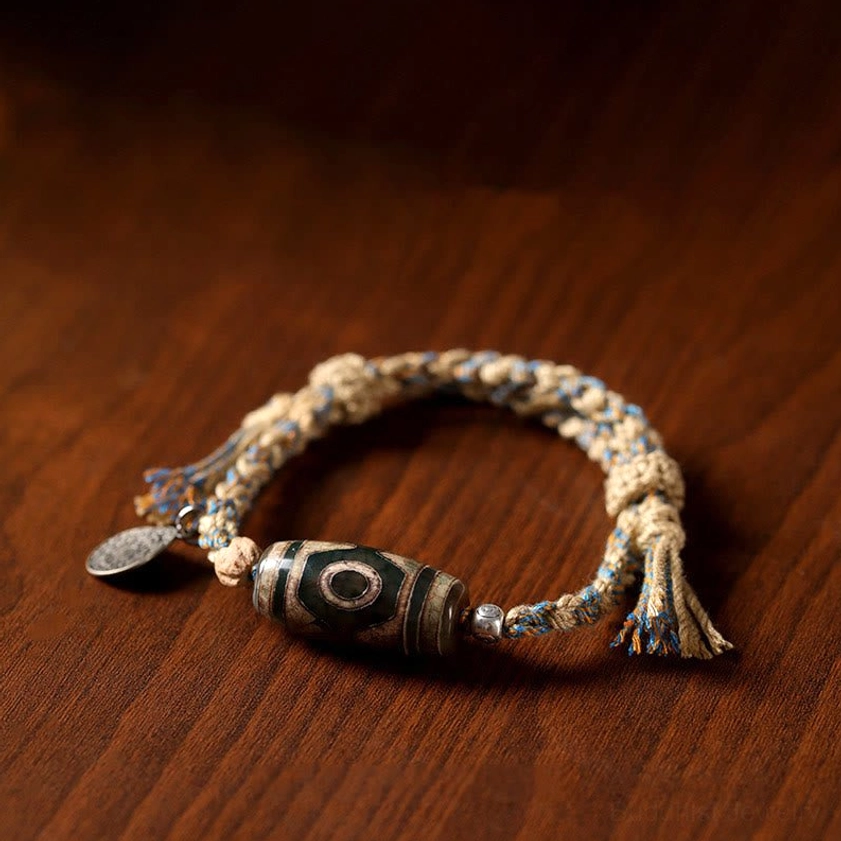 Three-eye Dzi bead bracelet flat lay on wooden table with metal tag and tassel ends, authentic handcrafted jewelry
