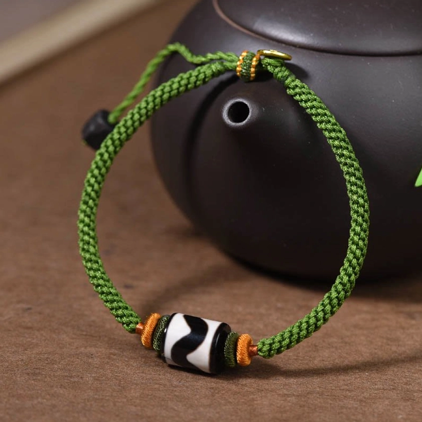 Three-quarter side view of green rope dzi bead bracelet against ceramic teapot background