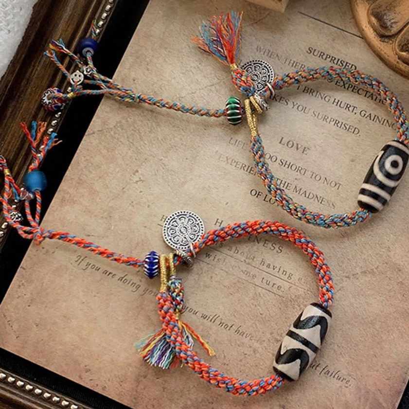 Two handmade Nine-Eye Dzi Bead protection bracelets with mixed-color woven ropes, silver charms, and black-white dzi beads on vintage display