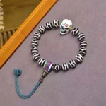 Handpainted dzi bead corpse forest lord protection bracelet with skull charm, blue cord, and striped beads displayed on wooden frame