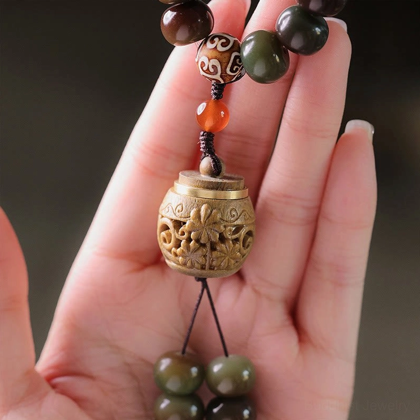 Dzi bead mala pendant detail with red agate spacer, carved wooden container, and green sandalwood tassel, finger-held close-up