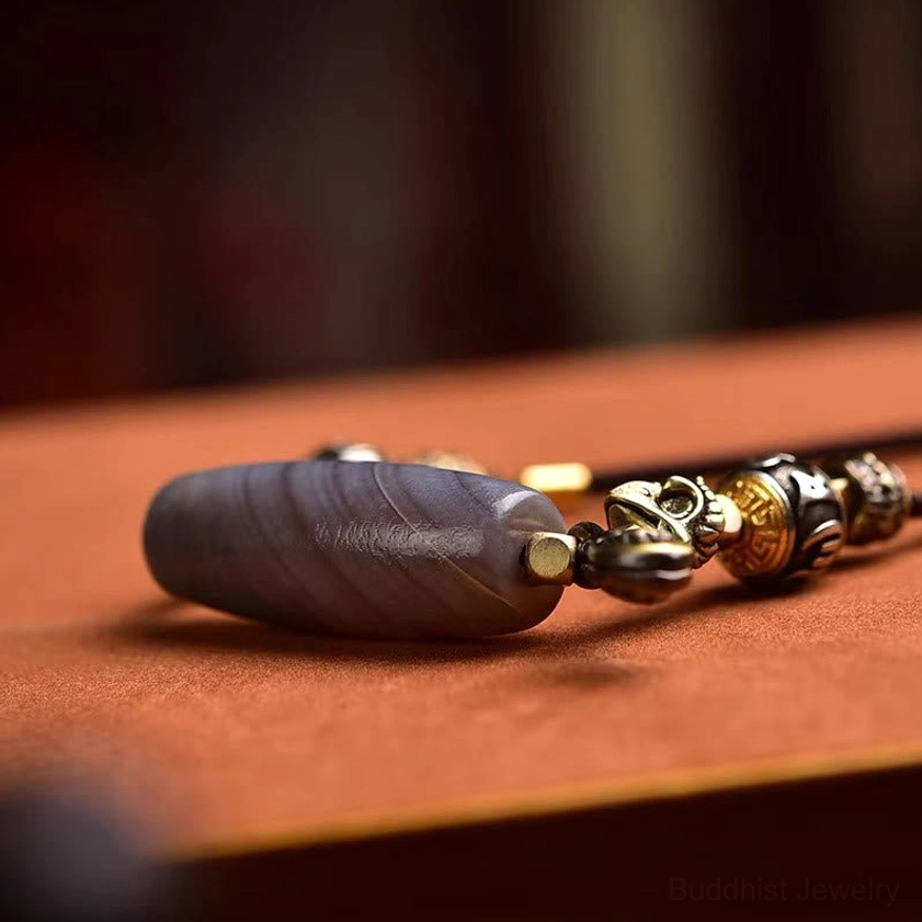 Buddhist necklace detail showing banded grey agate Dzi bead with mixed metal spacers and small charm pendant on brown wood