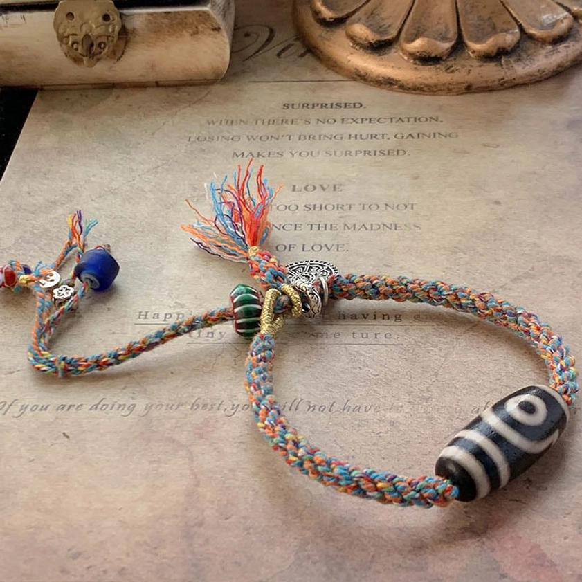 Nine-Eye Dzi Bead Buddhist protection bracelet with multi-color braided rope, silver charm, blue bead, and hand-painted dzi bead on vintage paper
