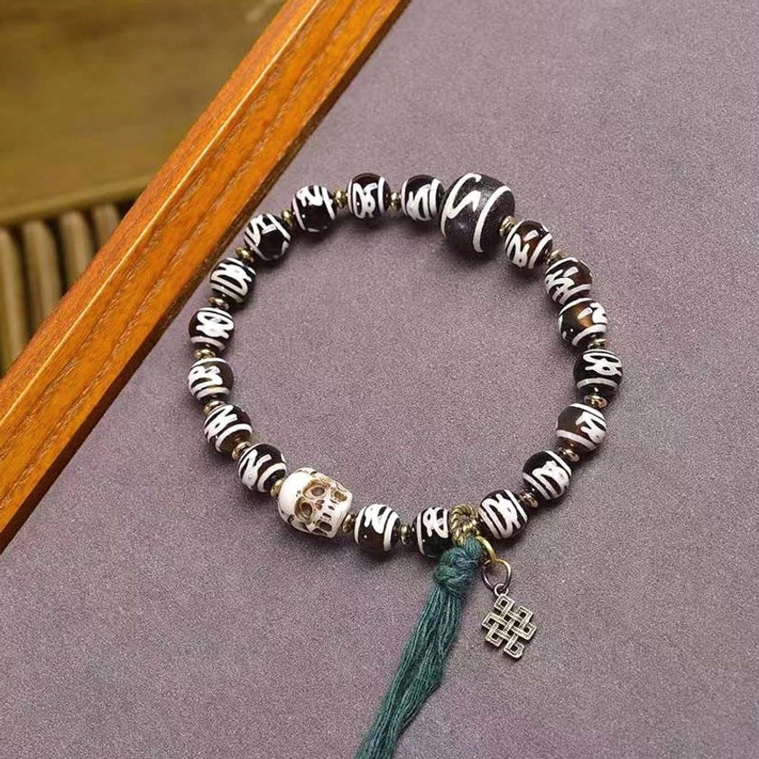 Dzi bead skull bracelet with black and white patterned beads, resin skull accent, and green tassel on purple fabric background