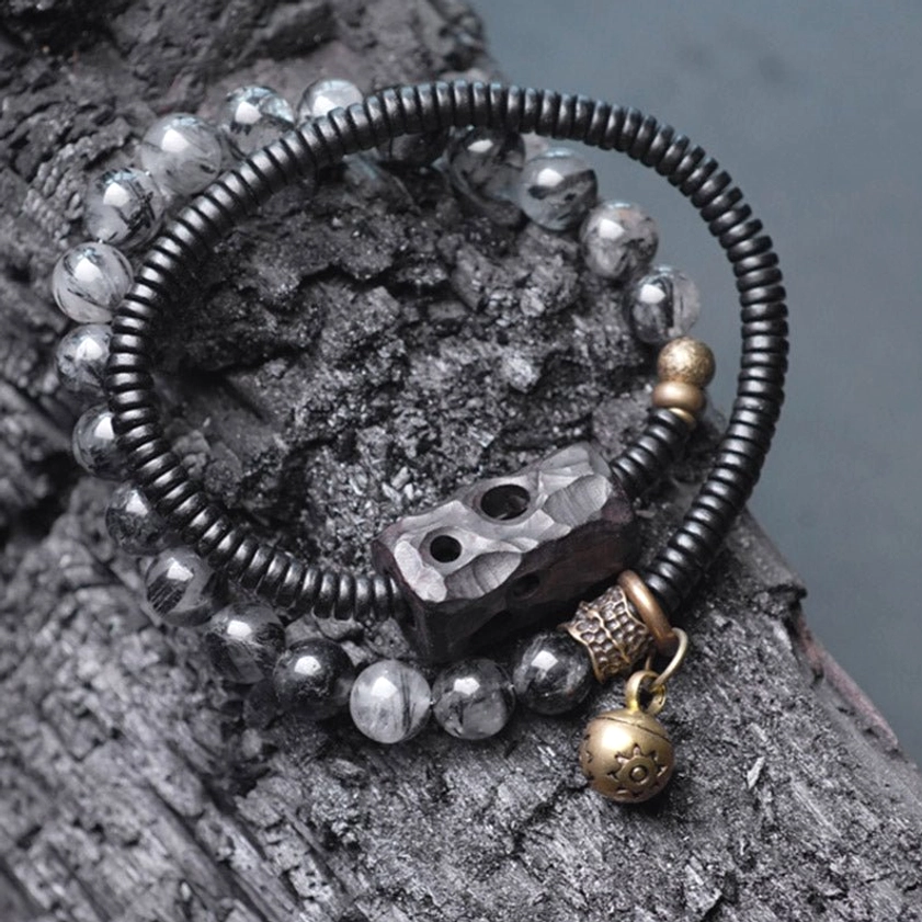 Double-layer beaded bracelet with hollow carved ebony wood bead and brass bell pendant on charcoal textured surface