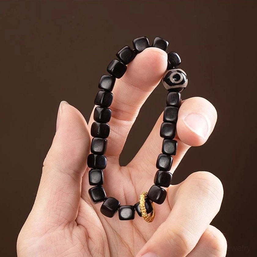 Hand-held close-up view of ebony wood dzi bead bracelet with nine-eye dzi bead, cubic beads, and gold metal ring on dark background