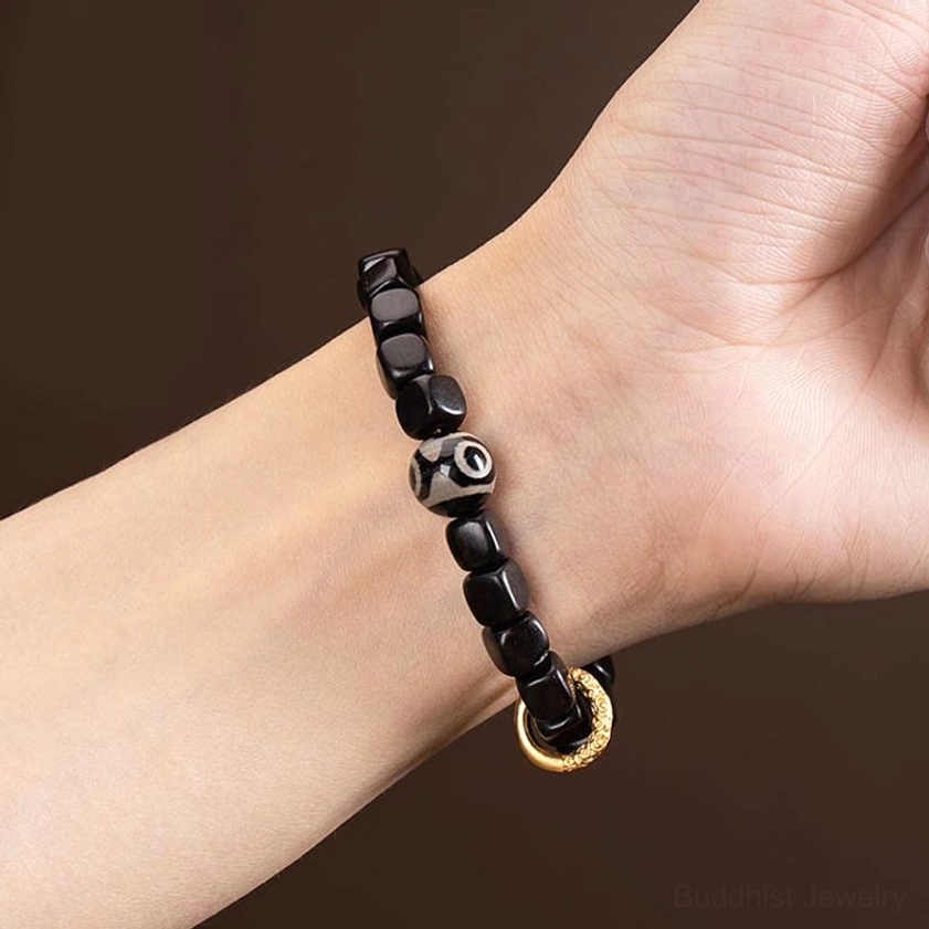 Ebony wood dzi bead bracelet worn on wrist showing cubic dark wood beads, nine-eye dzi design, and gold metal ring on black background, daily wear style