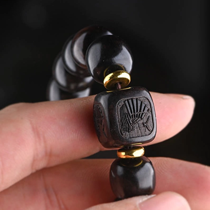 Close-up detail of handcrafted ebony wood barrel beads with engraved square charm and gold spacer rings held on fingertips