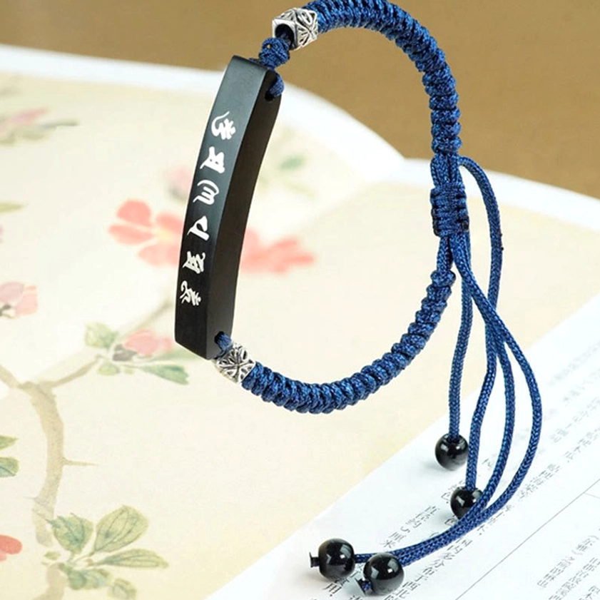 Side angle view of curved ebony wood Buddhist protection bracelet with multiple tassel adjusters and silver-inlaid Six True Words mantra tag on floral print paper background