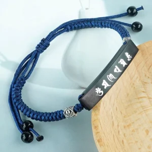 Ebony wood Om Mani Padme Hum Buddhist protection bracelet with silver inlay mantra tag, navy blue braided cord, and black agate adjusters displayed on wooden coaster with white ceramic decoration