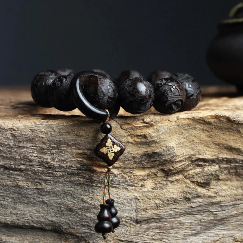 Side profile of hand-carved ebony bracelet showing diamond-shaped gilt charm and double gourd tassel on rough wooden display surface