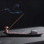 Handcrafted purple clay fisherman boat incense burner with burning incense stick, side three-quarter view showing traditional Buddhist meditation holder design
