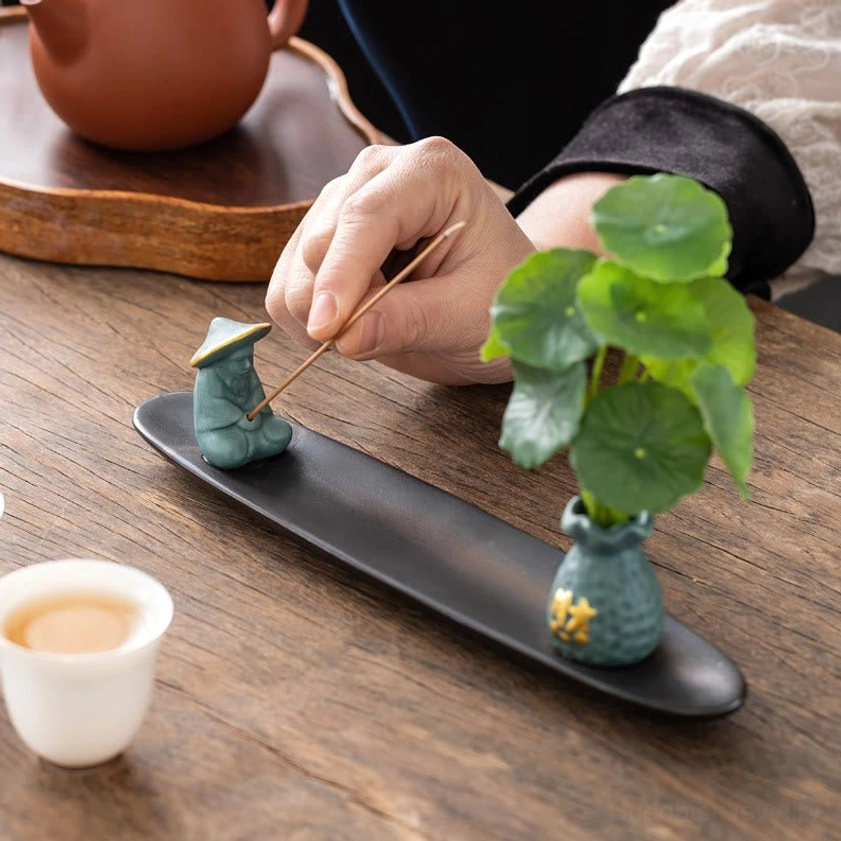 Close-up hand inserting incense stick into teal Fisherman ceramic Buddhist incense burner with figurine and vase