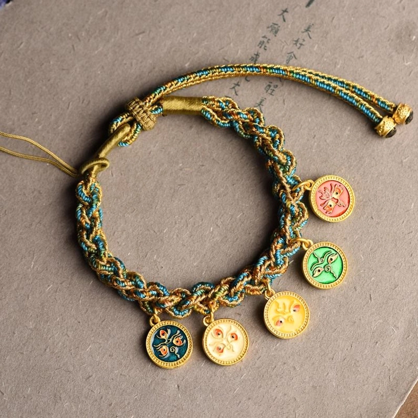 Five Gods of Wealth Buddhist string bracelet with blue-green-gold woven cord and four enamel decorative pendants on grey fabric backdrop