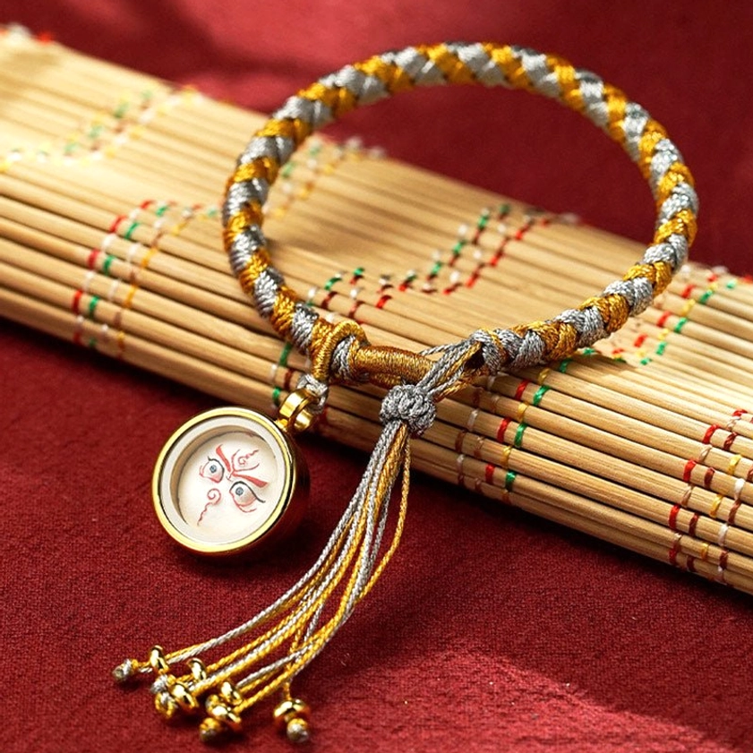 Five Gods of Wealth woven rope bracelet featuring gold-brown braid, white face amulet charm, and hanging tassel end, Buddhist prosperity piece