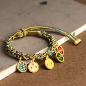 Five Gods of Wealth Buddhist bracelet overhead flat lay view with blue-gold woven rope and four enamel gold deity charms on wooden desk with stationery