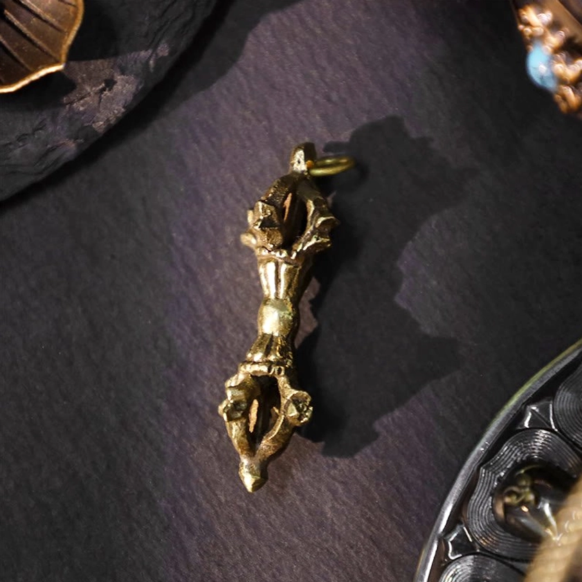 Single prong vajra pendant in close-up side view showing carved religious details, hanging loop, and aged brass finish on dark fabric