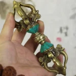 Handcrafted five-prong vajra dorje held in hand showing intricate turquoise inlays and carved scroll details against calligraphy background