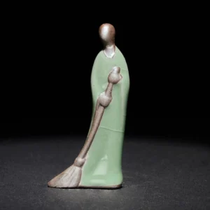 Formless Buddha sweeping monk ceramic statue left profile view with celadon green glaze and bronze details on dark background