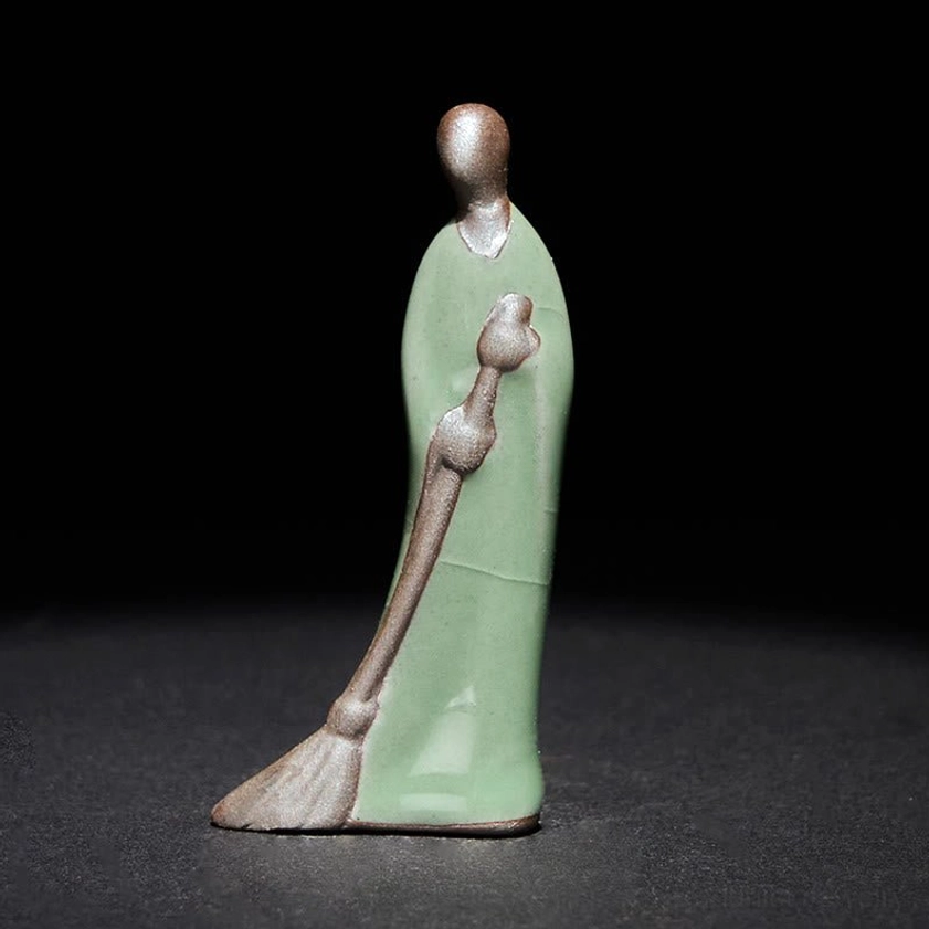 Formless Buddha sweeping monk ceramic statue left profile view with celadon green glaze and bronze details on dark background