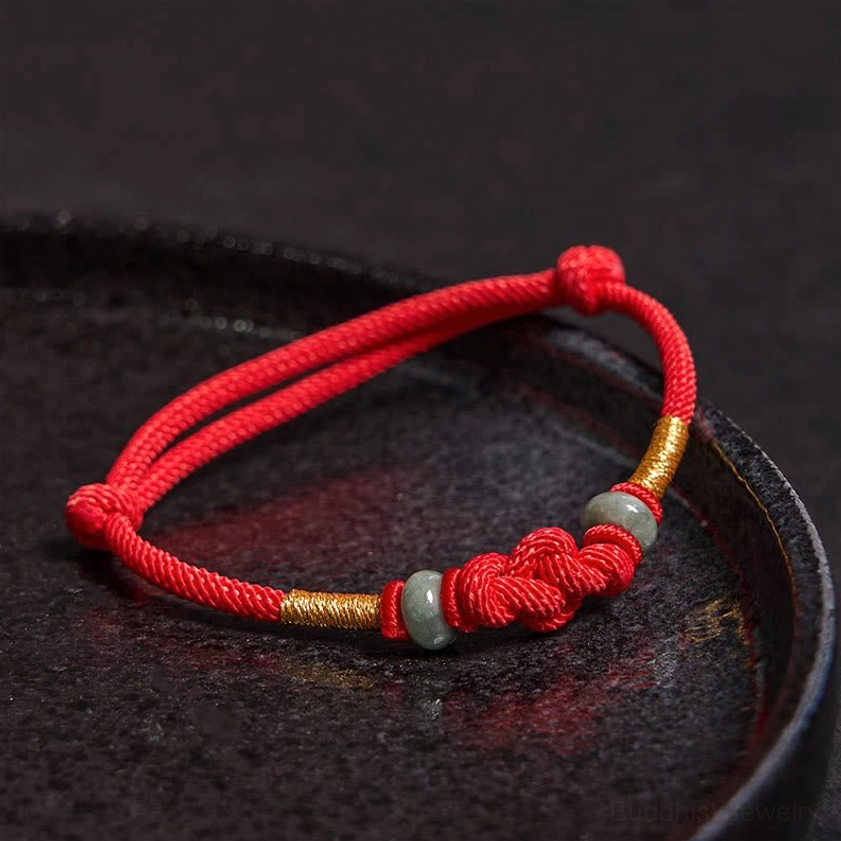 Buddhist fortune knot red string bracelet with symmetrical woven knots, gold thread segments, and light green jade beads overhead flat lay