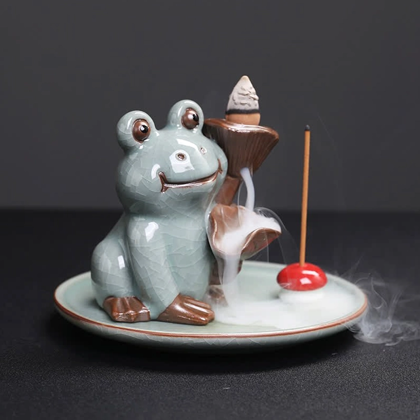 Buddhist frog incense burner with cascading incense smoke drifting over ceramic tray, celadon ceramic with brown decorative accents and left-side perspective