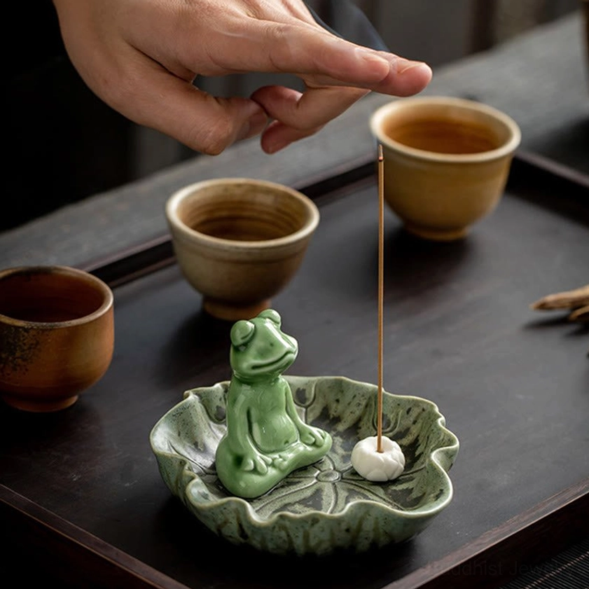 Hand adjusting incense stick in frog ceramic burner with visible aromatic smoke rising