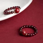 Garnet cinnabar PiXiu Om Mani Padme Hum Buddhist ring displayed on ceramic plate with wine-red background, showcasing burgundy beads, gold spacers, and hand-carved cinnabar charms