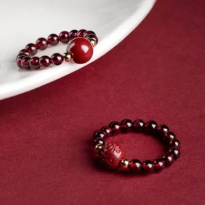 Garnet cinnabar PiXiu Om Mani Padme Hum Buddhist ring displayed on ceramic plate with wine-red background, showcasing burgundy beads, gold spacers, and hand-carved cinnabar charms