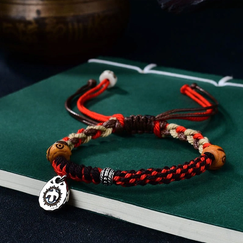 Hand-woven Garuda red string bracelet with braided design, metal tag, and adjustable knot closure on green book background
