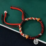 Red Garuda protection string bracelet with hand-woven tri-color cord, wooden beads, and Tibetan silver charm on dark display surface