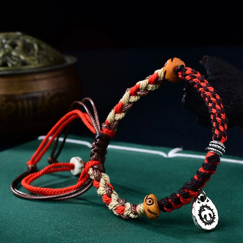 Buddhist protection string bracelet side angle showing red-brown woven rope, wooden beads, metal Garuda pendant, and adjustable straps